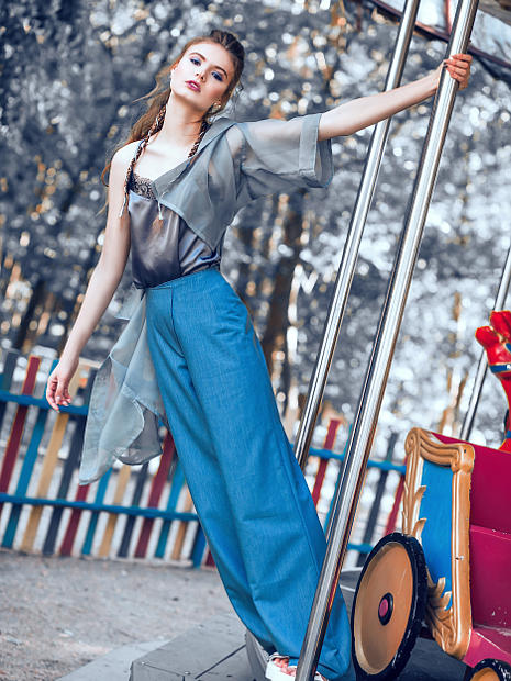 На фотографии девушка блондинка фото модель, которая стоит возле карусели и держится за никилированную трубу. Одета в темно синие брюки и серую блузку