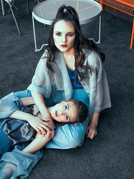 На фотографии две девушки модели, они на полу одна из них сидит а вторая лежит у ее ног на полу. Они держатся за руки.