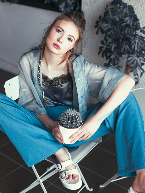 На фотографии девушка фото модель она сидит на стуле с кактусом в руках. Одета в шелковую прозрачную блузку и шелковые штаны.