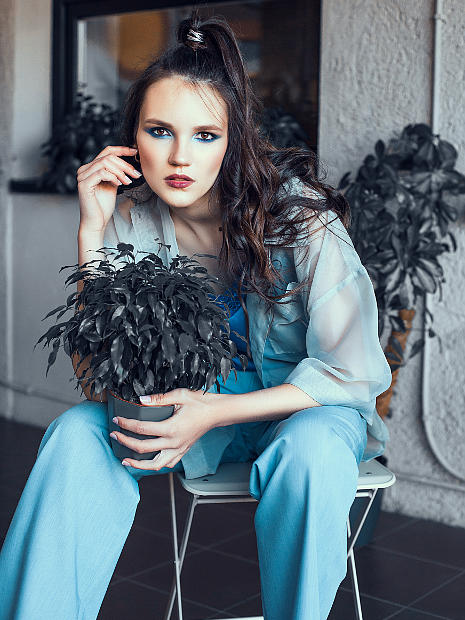 На фотографии девушка фото модель ярко накрашеная, она сидит на стуле у нее в руках горшок с цветком. Одета в шелковую прозрачную блузку и темно шелковые штаны.