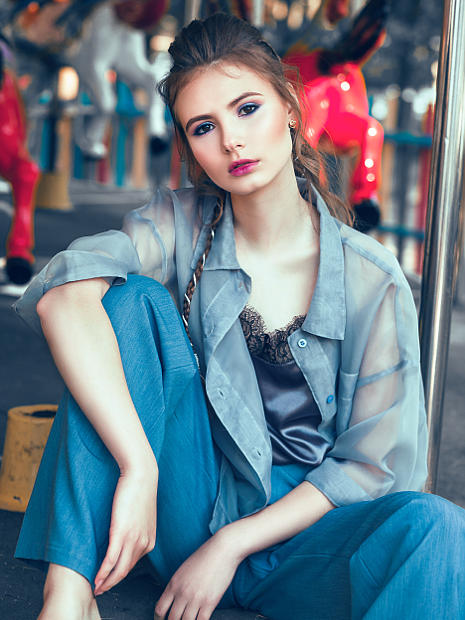 На фото фото модель двадцати лет. Она сидит на ступеньки возле карусели. В темно синих брючных штанах и серой блузке.