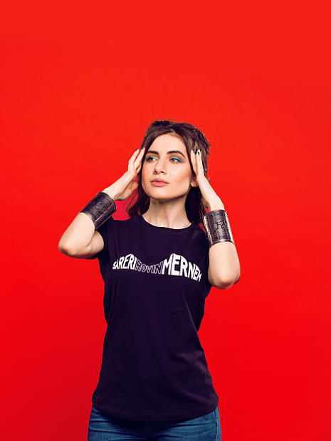 На фото девушка армянка в черной футболке с надписью Vuy Aman. Она взялась руками за голову у нее на руках широкие армянские браслеты.