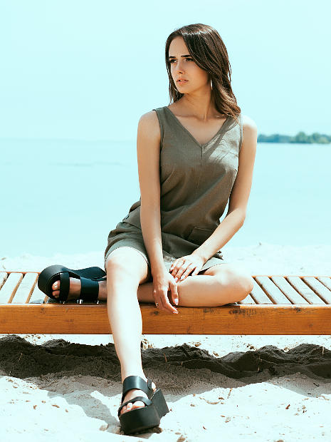 На фотографии девушка брюнетка модель рекламирует коричневый сарафан. Находятся она на пляже сидят на шезлонге
