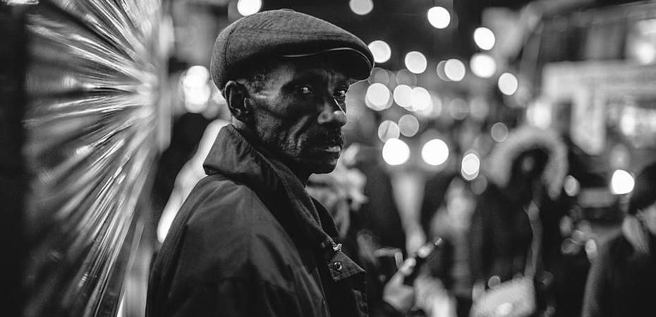 На фото темнокожий мужчина лет 50, в курточке и кепке на фоне ночного города. Профессиональный фотограф сделал спонтаное уличное фото.